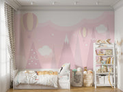 Pastel pink wall mural featuring hot air balloons, whimsical mountain peaks and soft clouds, decorating a cosy children’s bedroom with playful decor and a small bed.