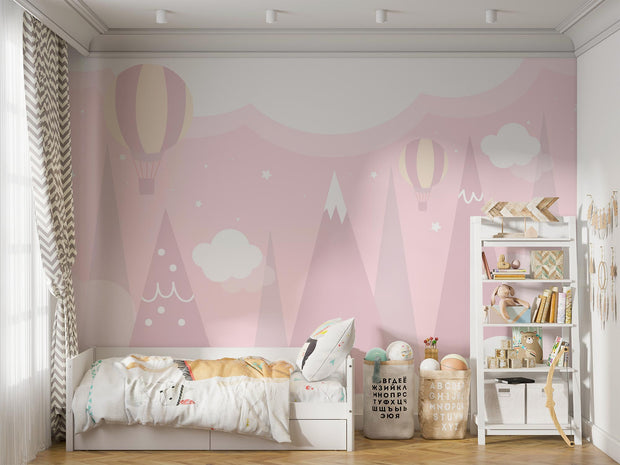 Pastel pink wall mural featuring hot air balloons, whimsical mountain peaks and soft clouds, decorating a cosy children’s bedroom with playful decor and a small bed.