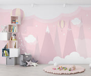 Pastel pink wall mural featuring hot air balloons, whimsical mountain peaks and soft clouds, decorating a cozy children’s playroom.