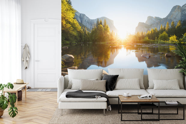 Mountain sunrise wall mural featuring a serene lake, evergreen trees, and soft morning light illuminating majestic mountains; decorating a living room.