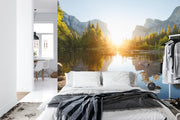 Mountain sunrise wall mural featuring a serene lake, evergreen trees, and soft morning light illuminating majestic mountains; decorating a bedroom.